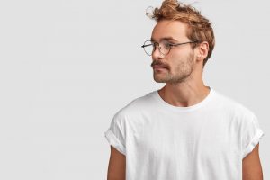 Profile shot of serious stylish hipster looks aside with confident expression, turns head aside, looks at something into distance, wears round spectacles, isolated over white background, blank space